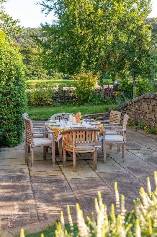 View of garden table, wine, chairs with cushions, trees, flowers, herbs and bench on patio