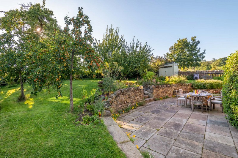 View of patio, lawn with apple trees bearing fruit, green shed, wheelbarrow and laid garden table with chairs and cushions