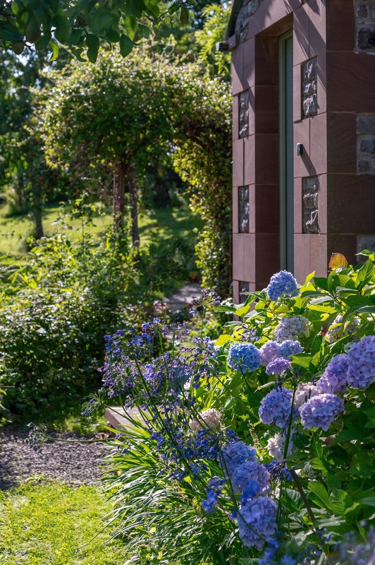 View of open front door and stone front of house with hydrangeas in bloom and garden in background