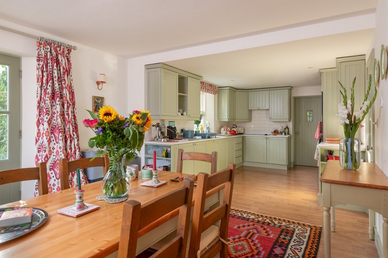 View of kitchen from front door, green cupboards, Linwood fabric, wood chairs and table with flowers and vibrant Kilim rug