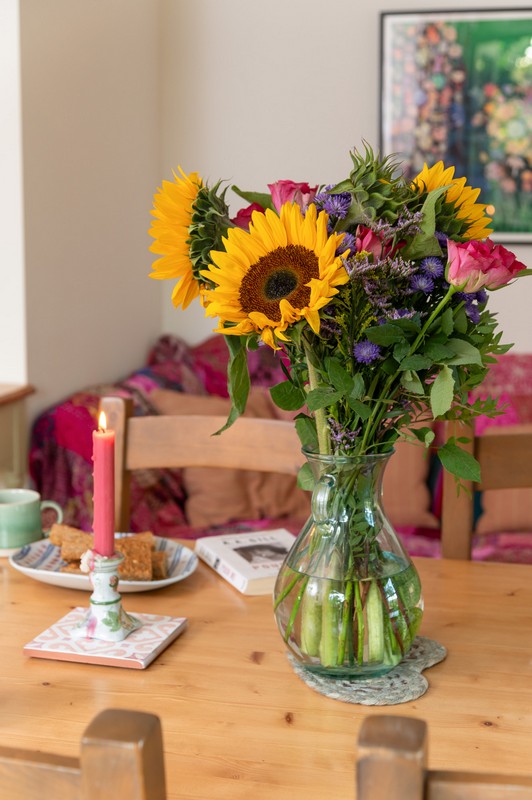 Flowers on kitchen table with lit candle, flapjacks, book tea, painting and sofa