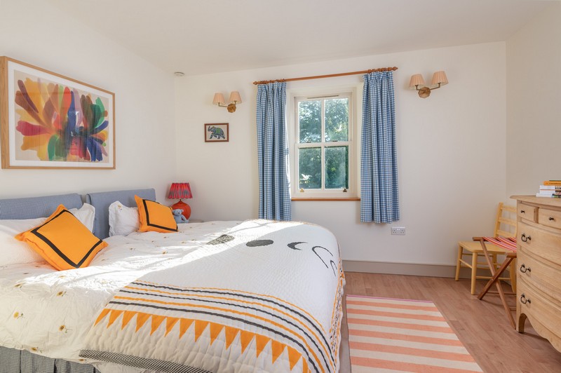 Second bedroom with blue gingham curtains and vibrant orange bedcover, cushions and lamp, with bright paintings and suitcase rack