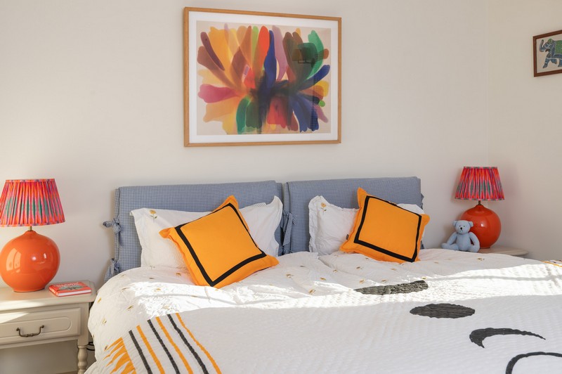 Second bedroom with vibrant orange cushions and lamps on matching bedside tables, with bright paintings and padded bedheads in blue gingham