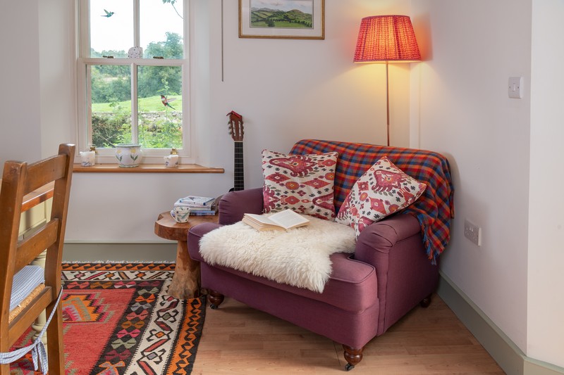 View of armchair in kitchen with brightly coloured cushions, sheepskin rug and blinds and lamp switched on